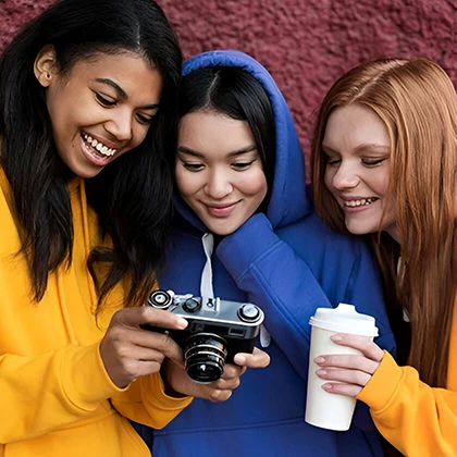Three girls wearing yellow hoodies pose with a camera, representing women's cultural impact through art and media.